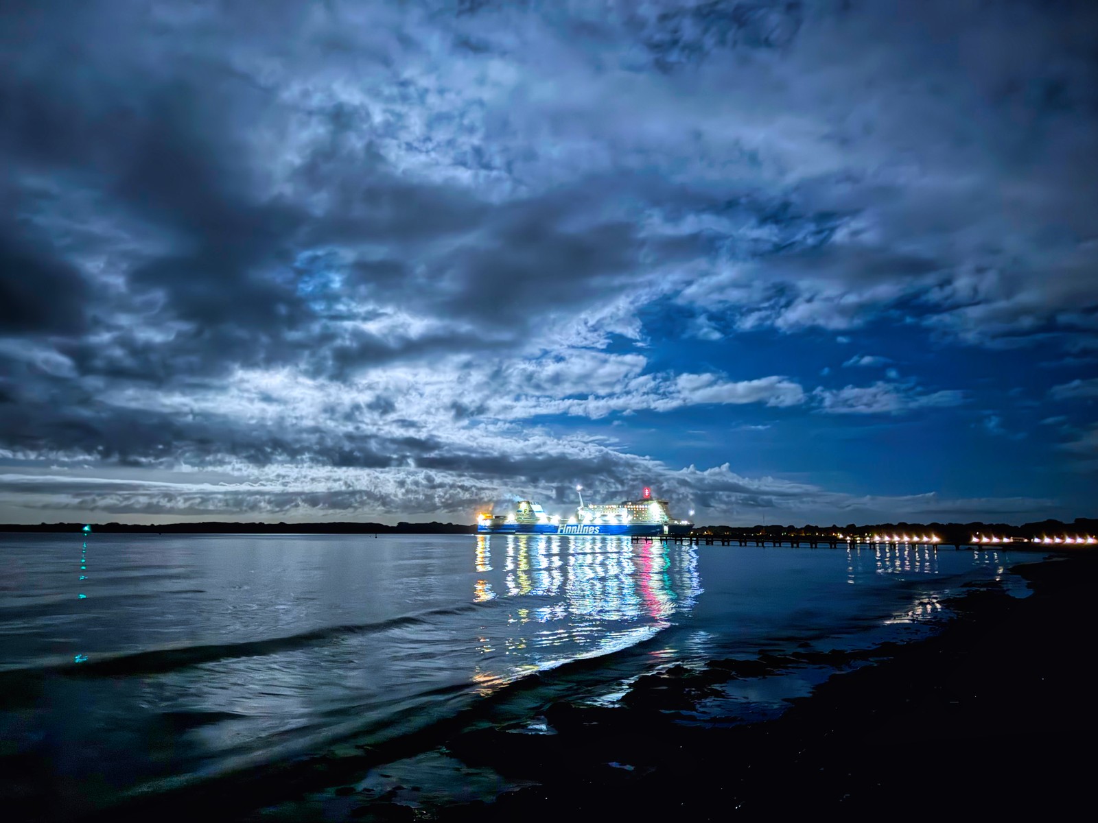 Das Bild zeigt eine Hafenszene bei Nacht. Im Vordergrund ist ein ruhiges, spiegelndes Gewässer zu sehen, in dem sich die Lichter der Schiffe und der Uferpromenade widerspiegeln. Am Horizont ist eine Reihe von beleuchteten Schiffen zu erkennen, die eine imposante Silhouette vor dem bewölkten Nachthimmel bilden. Die Stimmung ist mystisch und atmosphärisch, mit den Kontrasten zwischen Licht und Schatten, Wasser und Himmel.
