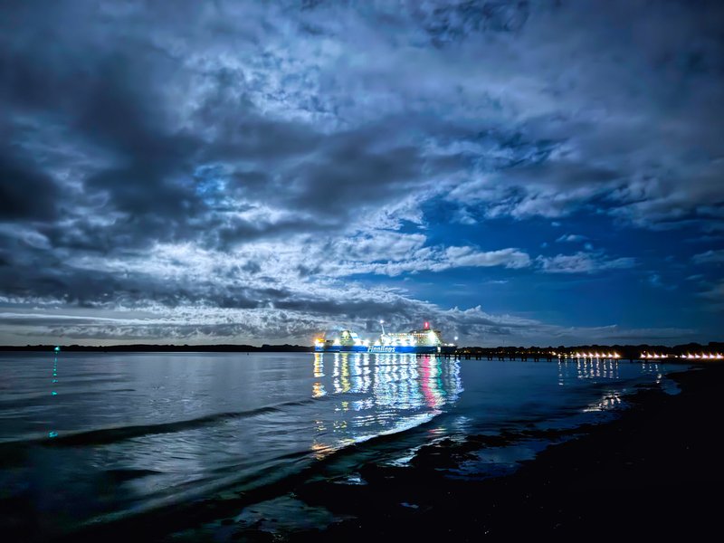 Das Bild zeigt eine Hafenszene bei Nacht. Im Vordergrund ist ein ruhiges, spiegelndes Gewässer zu sehen, in dem sich die Lichter der Schiffe und der Uferpromenade widerspiegeln. Am Horizont ist eine Reihe von beleuchteten Schiffen zu erkennen, die eine imposante Silhouette vor dem bewölkten Nachthimmel bilden. Die Stimmung ist mystisch und atmosphärisch, mit den Kontrasten zwischen Licht und Schatten, Wasser und Himmel.