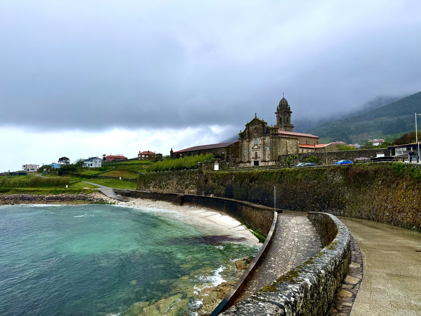 Das Bild zeigt eine malerische Küstenlandschaft. Im Vordergrund befindet sich eine steinerne Küstenmauer, die sich entlang des Wassers erstreckt. Dahinter erhebt sich eine barocke Kirche mit einem markanten Turm. Die Umgebung ist von sanft bewachsenen Hügeln und Gebäuden geprägt, die in einen nebligen, mystischen Dunst gehüllt sind. Insgesamt vermittelt das Bild eine ruhige, romantische Atmosphäre.