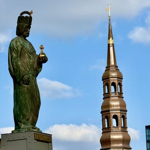 In dieser Aufnahme sehen wir eine große, grüne Statue eines bärtigen Mannes, der eine Art Krone trägt und ein Kreuz in seiner Hand hält. Im Hintergrund erhebt sich ein goldener, spitzer Kirchturm mit einer Kuppel. Der Kontrast zwischen der monumentalen Statue und dem eleganten Kirchenturm, der vor einem blauen Himmel mit weißen Wolken steht, schafft eine stimmungsvolle und historische Atmosphäre. Das Bild vermittelt einen Eindruck von Würde, Tradition und religiöser Symbolik an einem öffentlichen Ort.