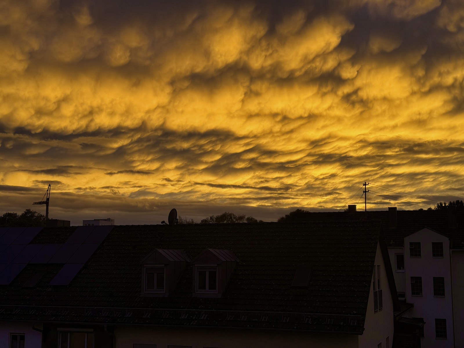 Das Bild zeigt eine dramatische Dämmerungsszene mit einem Himmel voller goldgelber und dunkelbrauner Wolken, die eine beeindruckende, beinahe majestätische Stimmung vermitteln. Silhouetten von Gebäuden und Strommasten zeichnen sich im Vordergrund ab, was dem Bild eine urbane Note verleiht. Die Beleuchtung und atmosphärische Stimmung lassen etwas Mystisches und Geheimnisvolles erahnen.