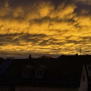 Das Bild zeigt eine dramatische Dämmerungsszene mit einem Himmel voller goldgelber und dunkelbrauner Wolken, die eine beeindruckende, beinahe majestätische Stimmung vermitteln. Silhouetten von Gebäuden und Strommasten zeichnen sich im Vordergrund ab, was dem Bild eine urbane Note verleiht. Die Beleuchtung und atmosphärische Stimmung lassen etwas Mystisches und Geheimnisvolles erahnen.