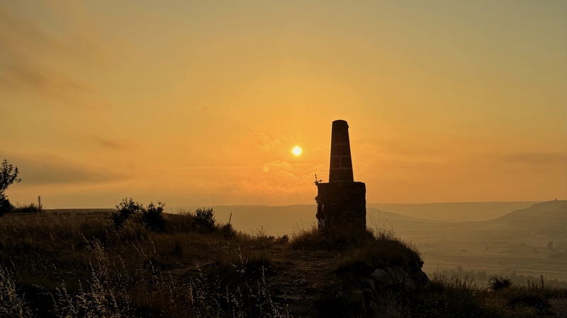 Ein steinernes Denkmal steht silhouettenhaft auf einem Hügel vor einem goldenen Sonnenaufgang. Die Sonne hängt tief über einer weiten Landschaft mit sanften Hügeln, während Gräser und Sträucher im Vordergrund im warmen Licht leuchten.