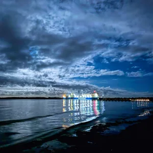 Das Bild zeigt eine Hafenszene bei Nacht. Im Vordergrund ist ein ruhiges, spiegelndes Gewässer zu sehen, in dem sich die Lichter der Schiffe und der Uferpromenade widerspiegeln. Am Horizont ist eine Reihe von beleuchteten Schiffen zu erkennen, die eine imposante Silhouette vor dem bewölkten Nachthimmel bilden. Die Stimmung ist mystisch und atmosphärisch, mit den Kontrasten zwischen Licht und Schatten, Wasser und Himmel.