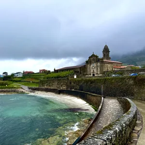 Das Bild zeigt eine malerische Küstenlandschaft. Im Vordergrund befindet sich eine steinerne Küstenmauer, die sich entlang des Wassers erstreckt. Dahinter erhebt sich eine barocke Kirche mit einem markanten Turm. Die Umgebung ist von sanft bewachsenen Hügeln und Gebäuden geprägt, die in einen nebligen, mystischen Dunst gehüllt sind. Insgesamt vermittelt das Bild eine ruhige, romantische Atmosphäre.