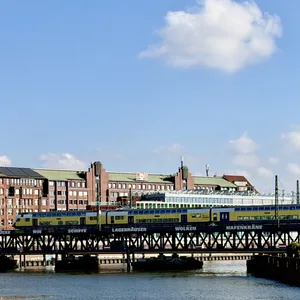 Das Bild zeigt eine urbane Landschaft mit mehrstöckigen Gebäuden, einer Eisenbahnbrücke und mehreren Zügen darauf. Die Gebäude haben einen historischen, industriellen Charakter mit einer Farbpalette von Rot, Grün und Gelb. Der Himmel ist hell und wolkig, was eine frische, sonnige Stimmung vermittelt. Insgesamt bietet das Bild einen lebendigen, visuellen Eindruck einer Stadt.
