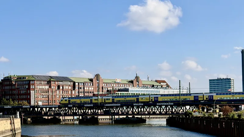 Das Bild zeigt eine urbane Landschaft mit mehrstöckigen Gebäuden, einer Eisenbahnbrücke und mehreren Zügen darauf. Die Gebäude haben einen historischen, industriellen Charakter mit einer Farbpalette von Rot, Grün und Gelb. Der Himmel ist hell und wolkig, was eine frische, sonnige Stimmung vermittelt. Insgesamt bietet das Bild einen lebendigen, visuellen Eindruck einer Stadt.