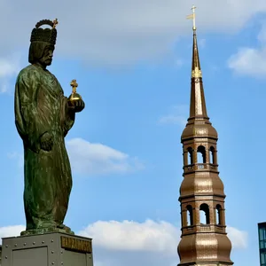 In dieser Aufnahme sehen wir eine große, grüne Statue eines bärtigen Mannes, der eine Art Krone trägt und ein Kreuz in seiner Hand hält. Im Hintergrund erhebt sich ein goldener, spitzer Kirchturm mit einer Kuppel. Der Kontrast zwischen der monumentalen Statue und dem eleganten Kirchenturm, der vor einem blauen Himmel mit weißen Wolken steht, schafft eine stimmungsvolle und historische Atmosphäre. Das Bild vermittelt einen Eindruck von Würde, Tradition und religiöser Symbolik an einem öffentlichen Ort.