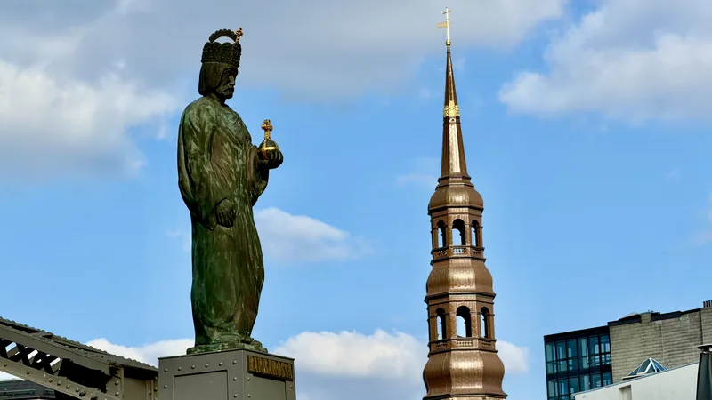 In dieser Aufnahme sehen wir eine große, grüne Statue eines bärtigen Mannes, der eine Art Krone trägt und ein Kreuz in seiner Hand hält. Im Hintergrund erhebt sich ein goldener, spitzer Kirchturm mit einer Kuppel. Der Kontrast zwischen der monumentalen Statue und dem eleganten Kirchenturm, der vor einem blauen Himmel mit weißen Wolken steht, schafft eine stimmungsvolle und historische Atmosphäre. Das Bild vermittelt einen Eindruck von Würde, Tradition und religiöser Symbolik an einem öffentlichen Ort.