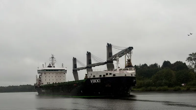Das Bild zeigt ein großes Frachtschiff namens "Viikki", das sich auf einem Fluss oder einer Wasserstraße befindet. Das Schiff ist dunkelgrün und hat auffällige Kräne und Strukturen an Bord. Hinter dem Schiff ist eine bewaldete Umgebung zu sehen, die eine eher düstere und ruhige Stimmung vermittelt. Das Bild wirkt insgesamt industriell und funktional.