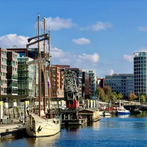 Das Bild zeigt einen Hafen mit einem historischen Segelschiff im Vordergrund. Dahinter erstreckt sich eine moderne Stadtlandschaft mit mehrstöckigen Gebäuden, die überwiegend aus Glas und Beton bestehen. Der Himmel ist blau mit einigen Wolken, was dem Bild eine freundliche und sonnige Stimmung verleiht. Insgesamt vermittelt das Bild einen Kontrast zwischen der traditionellen und modernen Architektur in einem urbanen Hafenumfeld.