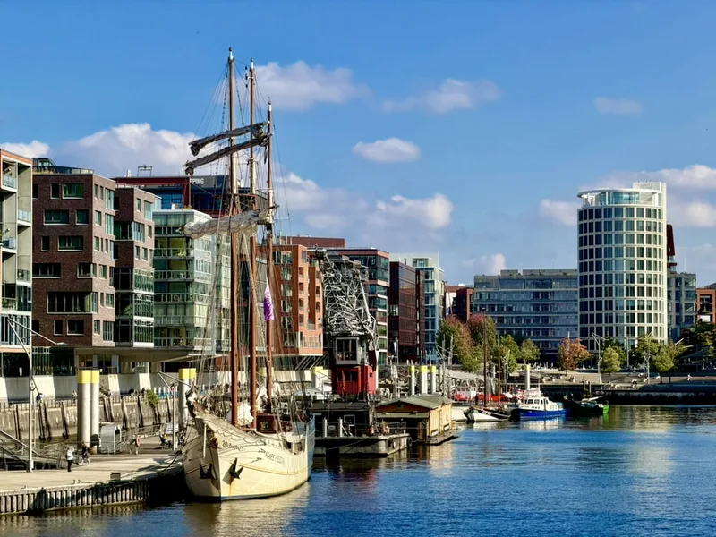 Das Bild zeigt einen Hafen mit einem historischen Segelschiff im Vordergrund. Dahinter erstreckt sich eine moderne Stadtlandschaft mit mehrstöckigen Gebäuden, die überwiegend aus Glas und Beton bestehen. Der Himmel ist blau mit einigen Wolken, was dem Bild eine freundliche und sonnige Stimmung verleiht. Insgesamt vermittelt das Bild einen Kontrast zwischen der traditionellen und modernen Architektur in einem urbanen Hafenumfeld.