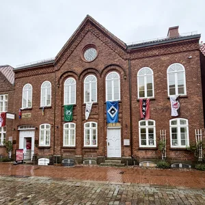 Das Bild zeigt ein zweistöckiges Ziegelsteingebäude mit großen Bogenfenstern und einem spitzen Dach. Das Gebäude hat einen historischen Charakter und vermittelt eine traditionelle europäische Architektur. Die Fassade ist mit Dekorationen verziert und es sind mehrere Eingangstüren zu sehen. Vor dem Gebäude befindet sich eine gepflasterte Fläche, die auf einen öffentlichen Platz oder eine Straße hinweist. Das Wetter erscheint bewölkt, was eine ruhige und gemäßigte Stimmung vermittelt.