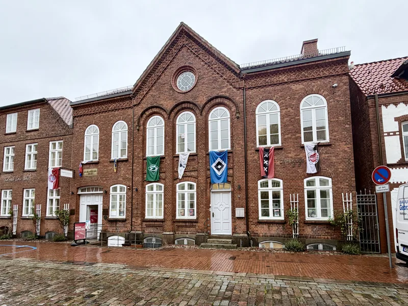 Das Bild zeigt ein zweistöckiges Ziegelsteingebäude mit großen Bogenfenstern und einem spitzen Dach. Das Gebäude hat einen historischen Charakter und vermittelt eine traditionelle europäische Architektur. Die Fassade ist mit Dekorationen verziert und es sind mehrere Eingangstüren zu sehen. Vor dem Gebäude befindet sich eine gepflasterte Fläche, die auf einen öffentlichen Platz oder eine Straße hinweist. Das Wetter erscheint bewölkt, was eine ruhige und gemäßigte Stimmung vermittelt.