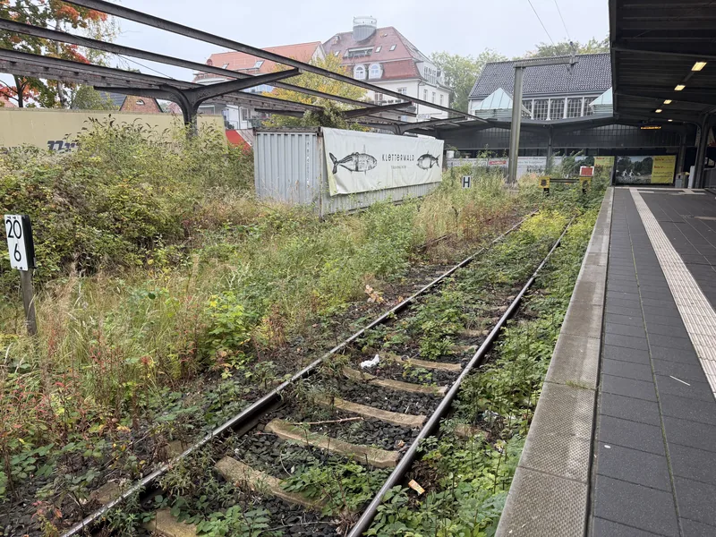 Das Bild zeigt eine Bahnstation oder Haltestelle mit verlassenen und verwilderten Gleisen. Der Bereich wirkt vernachlässigt, mit hohem Gras und Vegetation, die teilweise über die Gleise wuchert. An einer Wand ist ein Plakat mit einer Fischdarstellung angebracht. Die Umgebung vermittelt insgesamt einen etwas verlassenen und verwahrlosten Eindruck.