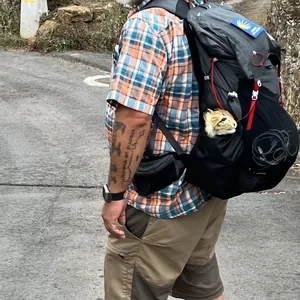 Das Bild zeigt einen Mann mit einem großen Rucksack auf dem Rücken, der einen gepflasterten Weg in einer naturnahen Umgebung entlanggeht. Der Mann trägt ein kariertes Hemd und Hosen in erdigen Farben. Der Rucksack hat eine rote Markierung und ein Luchs sitzt im Trinkfach. Die Umgebung wirkt ruhig und friedlich, mit Bäumen und Vegetation an den Seiten des Weges.