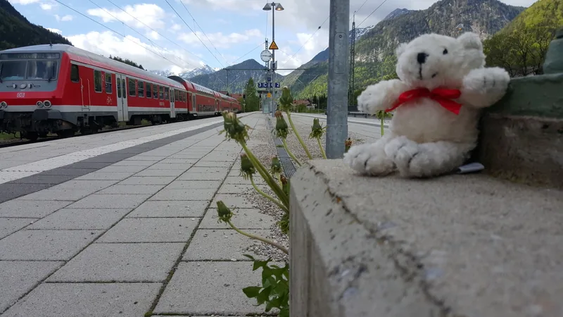Das Bild zeigt einen Bahnhof mit einer roten Regionalbahn auf dem Gleis. Der Bahnsteig ist aus grauem Stein und auf dem Geländer sitzt ein weicher, weißer Teddybär mit einer roten Schleife. Um den Bahnsteig herum sind die Berge und Wälder einer bergigen Landschaft zu sehen. Die Landschaft und der Zug vermitteln eine ruhige, idyllische Stimmung.