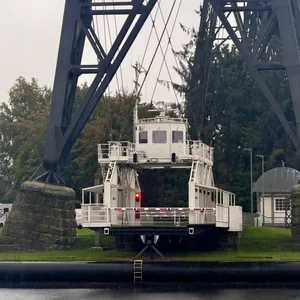 In diesem Bild sehen wir eine große, schwere Brücke, die über einen Fluss oder ein Gewässer führt. Unter der Brücke befindet sich ein Schwebefähre. Die Umgebung ist grün, mit einigen Gebäuden und Fahrzeugen in der Nähe. Die Stimmung erscheint düster und regnerisch, mit einem leicht melancholischen Gefühl.