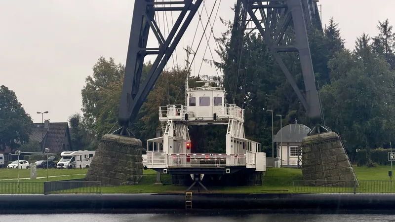 In diesem Bild sehen wir eine große, schwere Brücke, die über einen Fluss oder ein Gewässer führt. Unter der Brücke befindet sich ein Schwebefähre. Die Umgebung ist grün, mit einigen Gebäuden und Fahrzeugen in der Nähe. Die Stimmung erscheint düster und regnerisch, mit einem leicht melancholischen Gefühl.