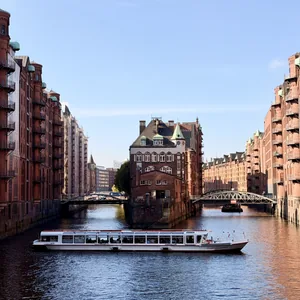 Das Bild zeigt einen malerischen Kanal in einer historischen Stadt, umgeben von prächtigen Backsteingebäuden mit Erkern und Türmen. Ein Ausflugsboot gleitet ruhig über das Wasser, während die ruhige Atmosphäre der alten Stadtarchitektur einen friedlichen und romantischen Eindruck vermittelt.