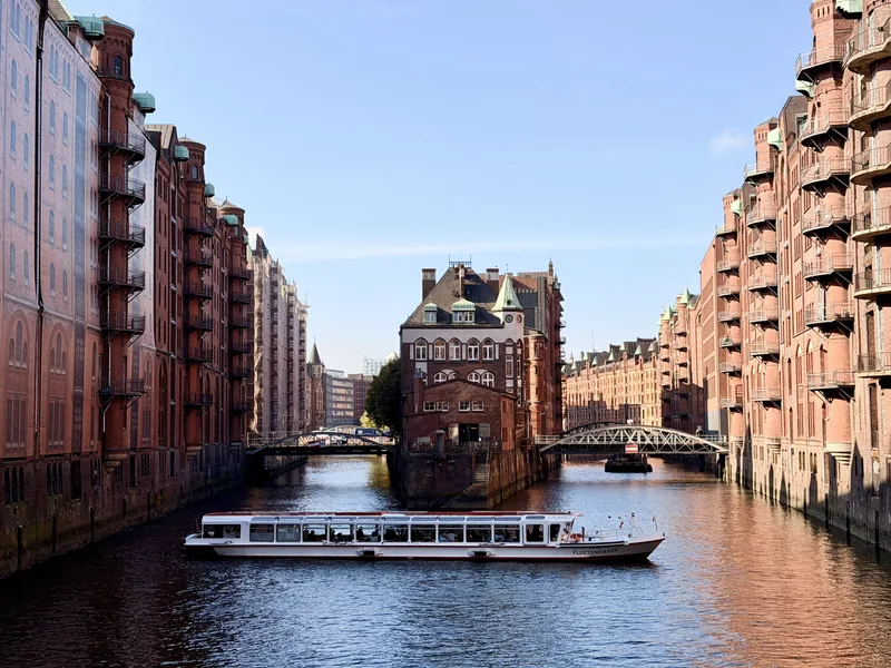 Das Bild zeigt einen malerischen Kanal in einer historischen Stadt, umgeben von prächtigen Backsteingebäuden mit Erkern und Türmen. Ein Ausflugsboot gleitet ruhig über das Wasser, während die ruhige Atmosphäre der alten Stadtarchitektur einen friedlichen und romantischen Eindruck vermittelt.