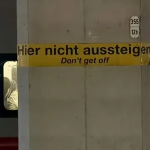 Das Bild zeigt einen Bahnsteig. Auf einer Säule ist der Text "Hier nicht aussteigen" auf Deutsch und Englisch ("Don't get off") zu sehen. Der Hintergrund ist dunkel und leer, was eine ernsthafte oder warnende Stimmung vermittelt. Es ist ein nüchternes, funktionales Bild, das eine klare Anweisung an Reisende übermittelt.