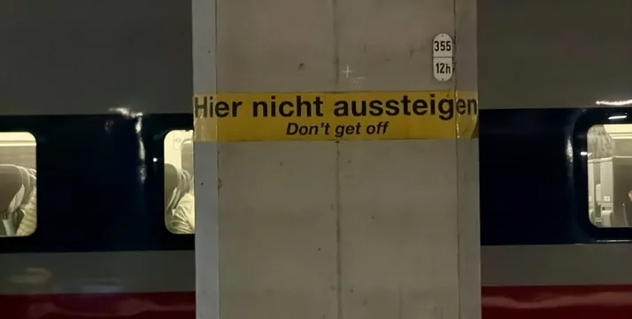 Das Bild zeigt einen Bahnsteig. Auf einer Säule ist der Text "Hier nicht aussteigen" auf Deutsch und Englisch ("Don't get off") zu sehen. Der Hintergrund ist dunkel und leer, was eine ernsthafte oder warnende Stimmung vermittelt. Es ist ein nüchternes, funktionales Bild, das eine klare Anweisung an Reisende übermittelt.